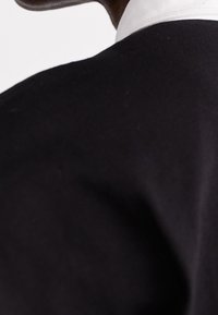 Schwarzes Kleidungsstück aus Stoff mit hohem Kragen. Der Kragen ist weiß und bildet einen Kontrast zum dunklen Material. Glatte Textur, zeigt Nähte und Form.