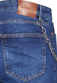 Jean en denim bleu avec une poche arrière cousue, une pièce en cuir portant l'inscription "2Y PREMIUM," et une chaîne argentée attachée à une boucle de ceinture.