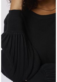 Femme portant un pull noir côtelé en maille avec des manches bouffantes plissées volumineuses et un collier fin en or, la main touchant ses cheveux.