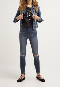 Giacca di jeans, t-shirt grafica nera, jeans blu strappati e ballerine nere con dettagli a strap. Outfit casual con texture contrastanti.
