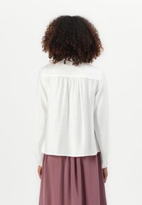 Kvinne med krøllete hår iført en løs hvit bluse med lange ermer og et mauve skjørt, stående med ryggen til kameraet.