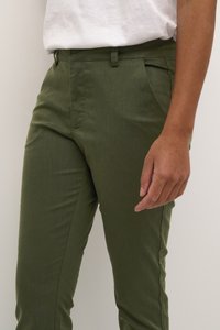 Olive green trousers made of textured fabric; slim fit with side pockets; paired with a plain white shirt.