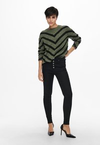 Schwarze, hochgeschnittene Skinny Jeans mit einem Fünf-Knopf-Verschluss, kombiniert mit einem grün-schwarz gestreiften Pullover im Zickzackmuster.