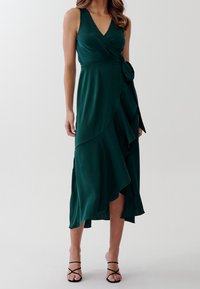 Kvinna som bär en ärmlös mörkgrön wrapklänning med volangig diagonalsöm och svarta remmar på högklackade sandaler mot en vit bakgrund.