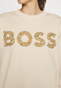 Pull beige avec "BOSS" écrit en grandes lettres pailletées dorées et argentées sur la poitrine, porté par une personne.