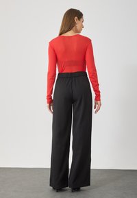 Un top lung roșu cu un textil neted, asortat cu pantaloni largi negri cu talie înaltă. Topul are un decolteu rotund și are un design ajustat.