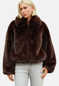 Femme portant une veste en fausse fourrure marron fonc�é avec un grand col et un jean gris clair, se tenant avec les mains détendues le long du corps.