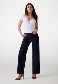 Top bianco a maniche corte con dettaglio a torsione, abbinato a jeans a vita alta e gamba larga blu scuro. Tacchi a punta marroni completano l'outfit.