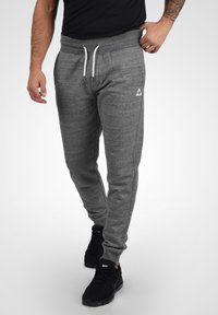 Pantalones deportivos grises con una cinturilla acanalada, cordón blanco, bolsillos laterales y un pequeño logo en la parte izquierda, combinados con zapatillas negras.