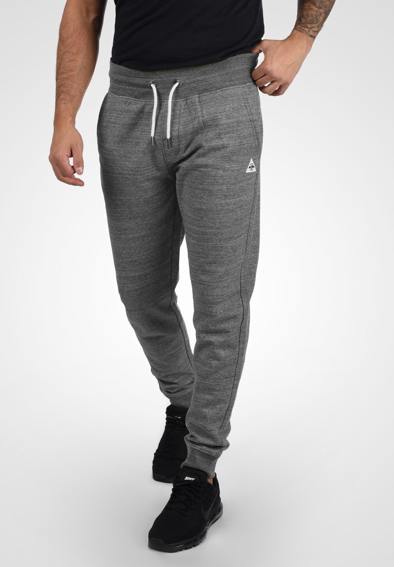 Pantalones deportivos grises con una cinturilla acanalada, cordón blanco, bolsillos laterales y un pequeño logo en la parte izquierda, combinados con zapatillas negras.