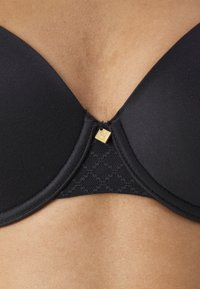 Schwarzer BH mit geformten Cups und Spitzenverzierung. Besitzt ein goldfarbenes Detail in der Mitte und eine glatte Stofftextur.