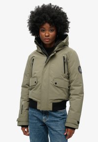 Zielona oliwka ocieplana kurtka z kapturem, z przednimi kieszeniami na zamek, czarnym ściągaczem u dołu oraz logo na ramionach.