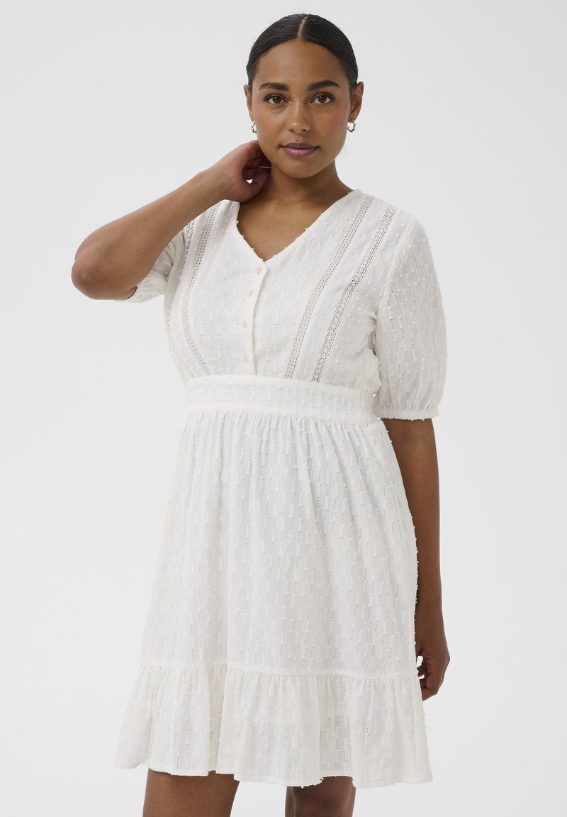 Femme aux cheveux foncés portant une robe blanche texturée mi-longue avec des boutons et des manches courtes bouffantes, debout devant un fond blanc.