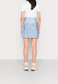 Nainen, jolla on päällään valkoinen t-paita, sinisiä pyörteisiä mini-hameita, valkoiset sukat ja oranssit Adidas-juoksukengät, seisoo selin tasaisella taustalla.