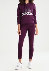 Sudadera morada con el logo blanco de "Adidas", combinada con leggings morados a juego. Zapatillas blancas completan el look atlético. Textura suave, diseño ajustado.