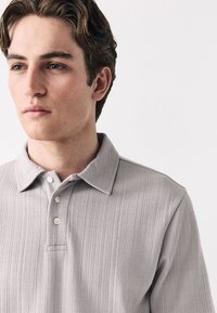 Polo grigio chiaro con tessuto strutturato, colletto e patta con tre bottoni. Presenta sottili strisce orizzontali per un interesse visivo.