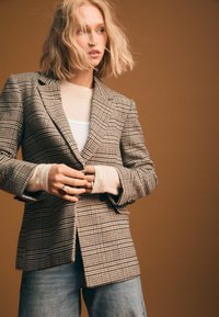 Blazer a quadri in tessuto marrone e crema con design su misura. Indossato sopra un morbido top beige e abbinato a pantaloni di jeans. Dettaglio con anello.