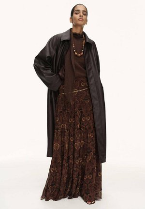 Longo vestito marrone con motivi intricati, abbinato a una maglietta marrone scuro e a un cappotto di pelle nero oversize. Accentuato con una collana di perline.