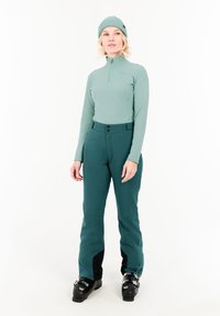Mintgroene lange mouw top met ritssluiting, gecombineerd met donkergroene waterdichte broek met een zwarte onderpaneel en zwarte skibotten.