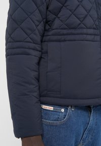 Veste matelassée bleu marine avec col montant, poignets côtelés et poche latérale. Associée à un jean bleu en denim et une étiquette Calvin Klein.