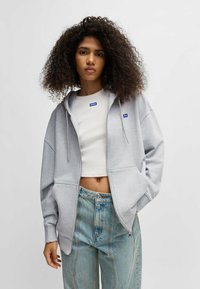 Light gray zip-up hoodie with a relaxed fit, two front pockets, and blue logo patches, paired with a white crop top and faded blue jeans.