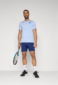 T-shirt de sport bleu clair à manches courtes avec logo noir, associé à un short marine, des tennis noirs et des chaussettes blanches, tenant une raquette de tennis.