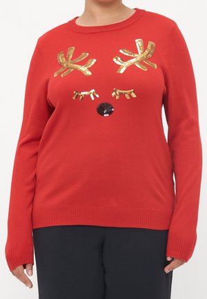 Czerwony sweter z złotymi cekinowymi porożami, zamkniętymi oczami i czarnym cekinowym nosem przypominającym renifera na torsie, noszony przez osobę w czarnych spodniach.