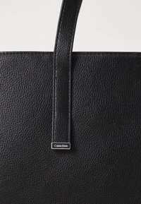 Czarna skórzana torba typu tote z wyraźnym paskiem i srebrnym akcentem z logo Calvin Klein. Prosty i elegancki design.