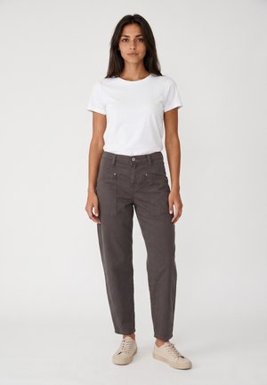 Femme debout portant un t-shirt blanc uni, un pantalon gris foncé avec de grandes poches avant et des baskets décontractées beiges sur fond blanc.