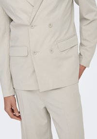 Chaqueta de traje beige claro de doble botonadura con solapas muescas, dos botones y bolsillos laterales. Fabricada en una tela suave y texturizada.
