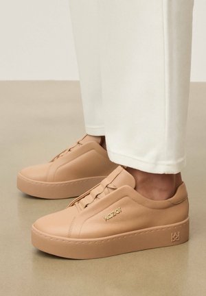 Bēšas ādas platformas kedas ar iespiestu “Kazar” logotipu sānos, nēsātas ar baltām biksēm, stāvot uz gludas grīdas.