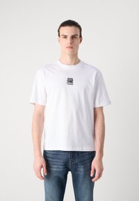 T-shirt en coton blanc à manches courtes, arborant un logo graphique noir sur la poitrine. La coupe est décontractée, avec un col rond.