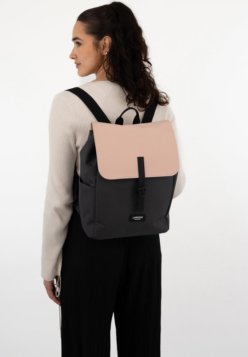 Zaino con un flap sintetico rosa e corpo in tessuto nero, spallacci regolabili e dettaglio in fibbia nera. Etichetta del brand visibile.