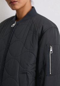 Gros plan sur une personne portant une veste bomber matelassée noire avec une fermeture éclair à l'avant et une poche zippée sur la manche supérieure.
