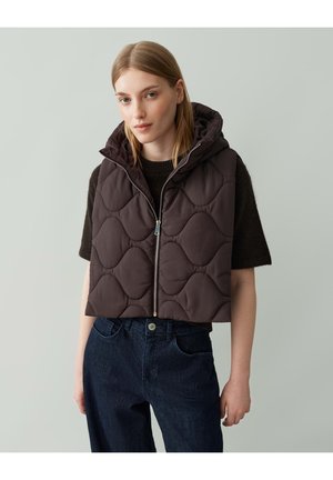 Brown padded vest with a hood, featuring a quilted design, zip closure, and layered over a dark sweater. Worn with dark denim pants.