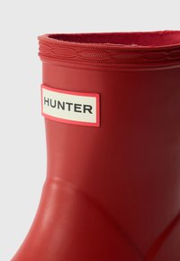 Rdeča gumijasta dežna čevlja s teksturirano zgornjim delom in ravnim peto. Ima belo etiketo s črnimi črkami "HUNTER" na strani.