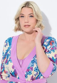 Bata con estampado floral que presenta patrones vibrantes en azul, rosa y verde sobre un fondo crema, con un cinturón lila sólido y mangas anchas.