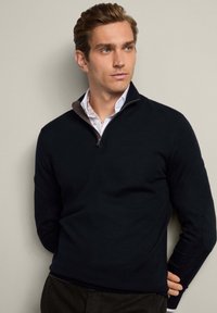 Marinblå stickad tröja med halvdolt dragkedja, med en texturerad brun foder. Långa ärmar med diskreta armbågsdetaljer, tillverkad i mjukt material.