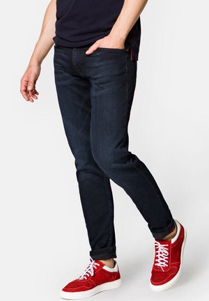 Ciemnoniebieskie dżinsy o slim fit, z podwiniętymi mankietami. Noszone z czerwonymi zamszowymi tenisówkami z białymi sznurówkami i białą podeszwą. Prosty, casualowy design.