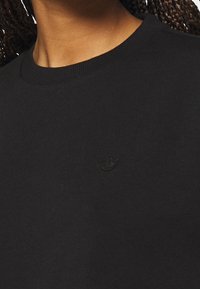 Närbild av en person som bär en svart rundhalsad tröja med en liten, diskret broderad logga på bröstet.