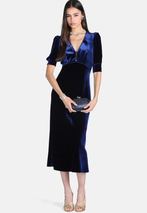 Femme portant une robe midi en velours bleu profond avec des manches courtes bouffantes, tenant une petite pochette noire, accessoirisée d’un collier, d’un bracelet et de boucles d’oreilles.
