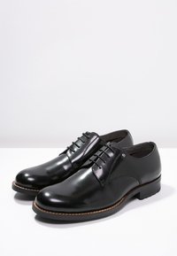 Chaussures habillées en cuir noir avec une finition lisse, à lacets et à coutures beiges. Présentent un bout arrondi et un logo discret sur le côté.