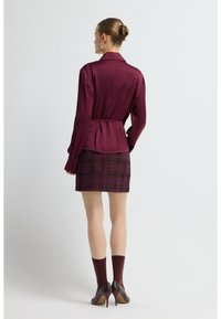 Bordeaux satin skjorte med bælte i taljen, parret med en ternet nederdel med teksturerede accenter, suppleret med mørke, hælede sko og strømper.