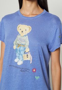 Modrá tričková košile s krátkým rukávem, na které je grafika medvěda v svetru a džínech, jak drží košík. Vyšívané detaily zahrnují srdce a květinu.