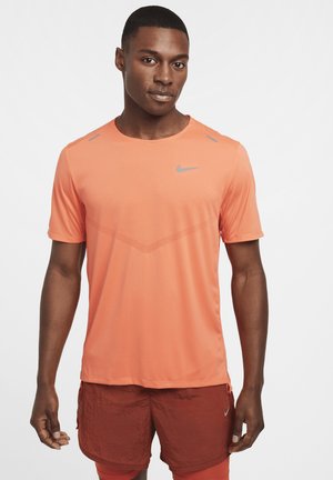 T-shirt sportivo arancione con maniche corte, collo rotondo, tessuto testurizzato, logo grigio sul petto, accenti a contrasto sulle spalle, abbinato a pantaloni corti rossi.