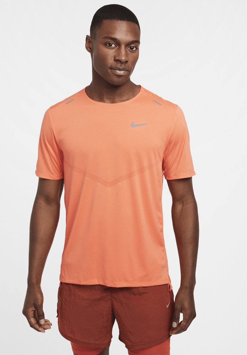 Camiseta deportiva naranja de manga corta, con cuello redondo, tejido texturizado, logo gris en el pecho, acentos en los hombros de color contrastante, combinada con pantalones cortos rojos.