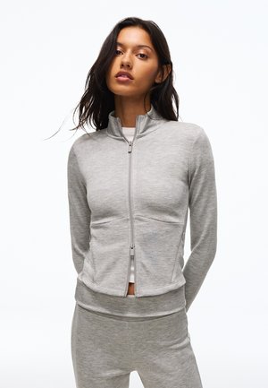 Femme aux longs cheveux foncés portant une veste zippée gris clair et un pantalon assorti sur un fond blanc uni.