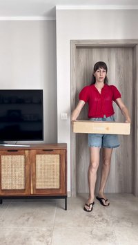 Cardboard box held in both hands, light brown color with white branding. Background features a wooden door and a wooden cabinet with wicker accents.