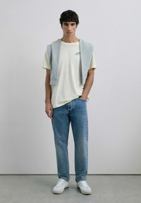 Maglietta gialla chiara con ricami blu, felpa grigia drappeggiata sulle spalle, jeans di denim blu e sneaker bianche. Modello in piedi contro uno sfondo neutro.