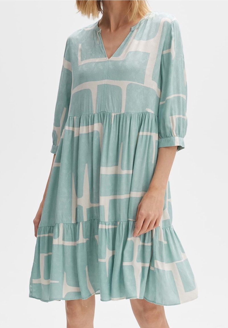 Robe ample bleu clair avec motif géométrique abstrait blanc, manches trois-quarts, encolure en V et jupe à volants, portée par une femme.